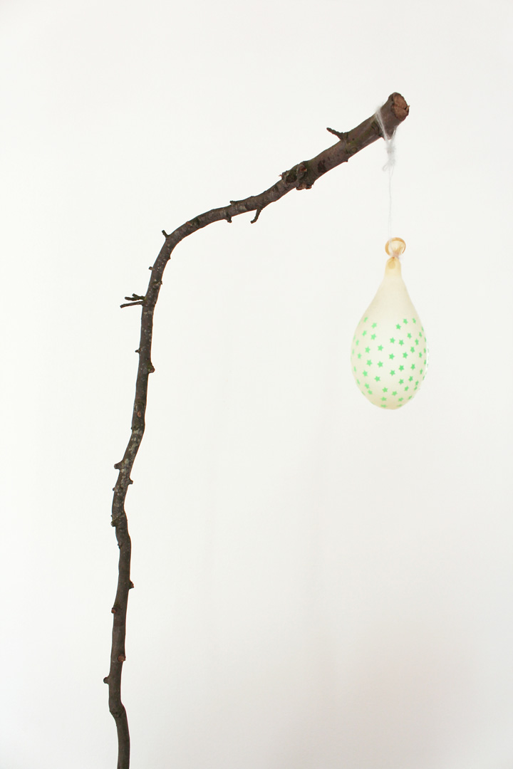 Vue d'exposition de l'installation intitulée " Aux corneilles". Focus resserré montrant une branche coudée à laquelle pend un ballon de baudruche étoilé de vert clair. Le ballon est à peine rempli d'air.