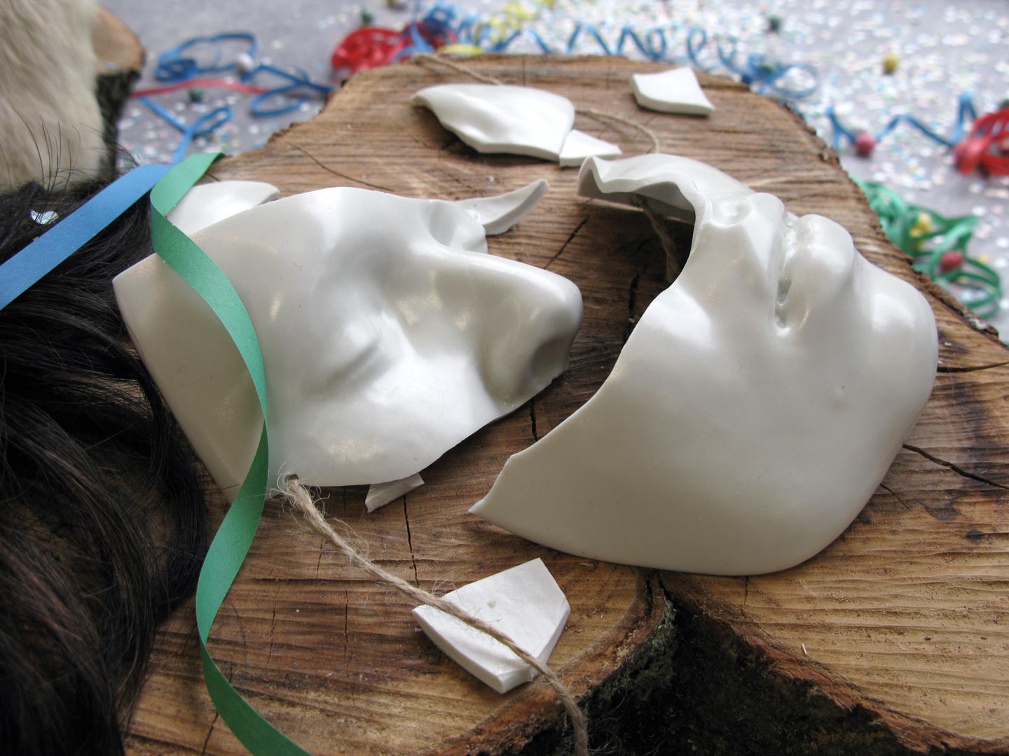 Vues de l'installation intitulée "Joyeuses Pétoches". Focus resserré sur le personnage morcelé du centre au masque cassé. Le personnage est déposé sur un tronc d'arbre. Son masque est en porcelaine émaillée blanche. Il est brisé.