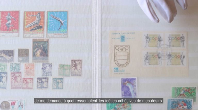 Arrêt sur image de la vidéo intitulée "Debout sous la langue". L'image présente une double page d'une album de philatélie partiellement remplie de timbres aux représentations dites "masculines" . En haut, à droite, l'extrémité d'une main gantée manipule la page. Une phrase en sous-titrage, centrée au bas, inscrit : "Je me demande à quoi ressemblent les icônes adhésives de mes désirs".