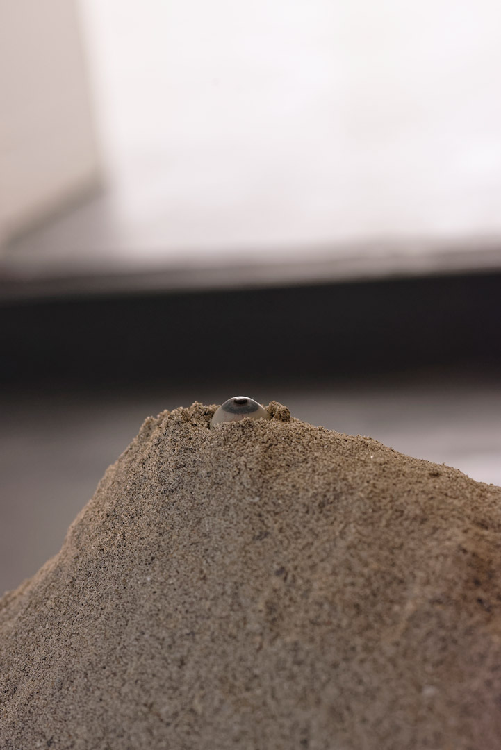 Vue de l'installation intitulée " Les tombeaux innocents". Focus sur un monticule de sable. Sur sa crête, un œil de verre hésite entre surgissement et enlisement.