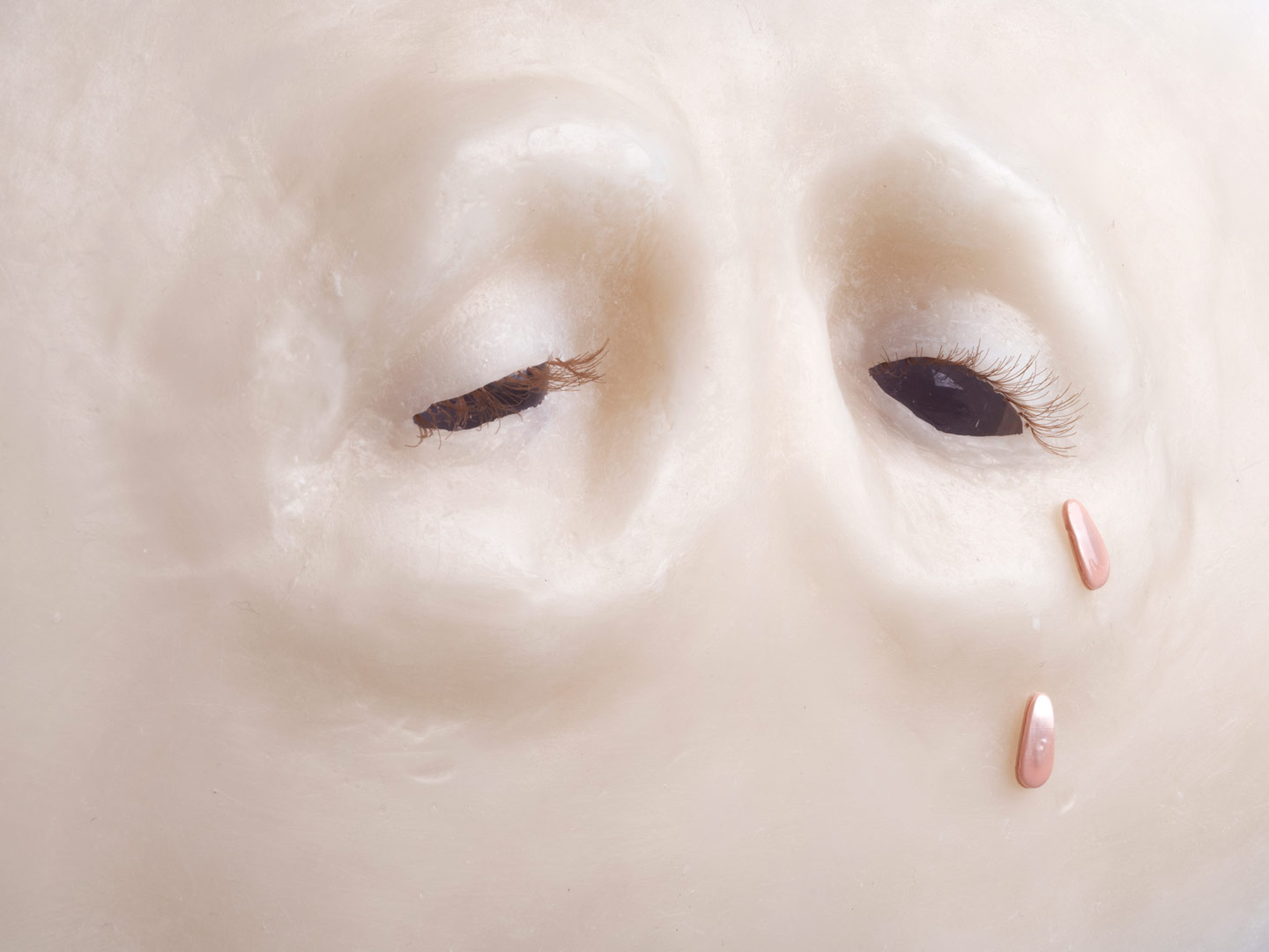 Fragment de l'installation Spleen Spring. Focus sur un regard en cire au nez et aux sourcils absents. Les yeux sont bordés de cils blonds implantés. Les globes oculaires, sont figurés par des sphères de verre transparentes qui laissent échapper deux larmes roses iridescentes.