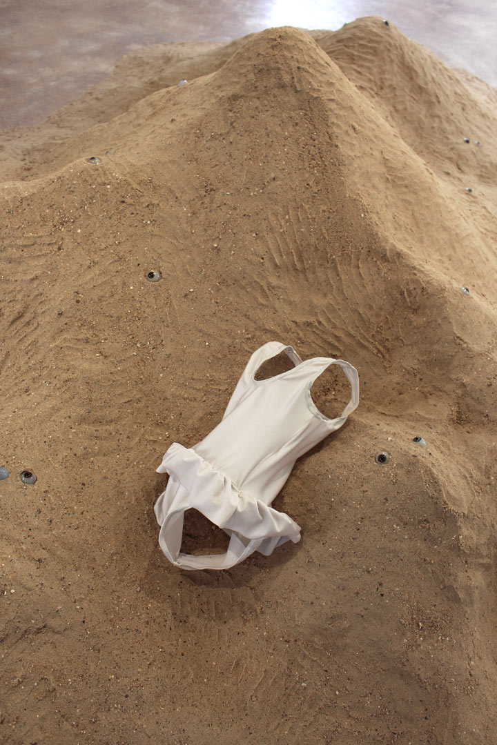 Vue de l'installation intitulée " Les tombeaux innocents". Focus resserré sur un maillot de bain déposé dans le sable. Il semble occupé mais pourtant vide. Des yeux de verre de différentes couleurs hésitent entre surgissement et enlisement. Ils semblent regarder dans différentes directions.
