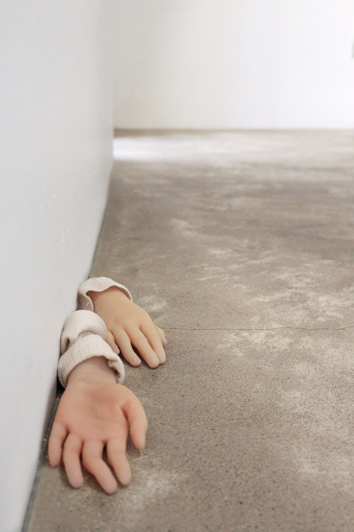 Vue d'exposition de l'installation intitulée "Les Vieux démons". Focus resserré sur une paire de mains déposées nonchalamment au sol. Des manches roses pales enlacent les poignets.