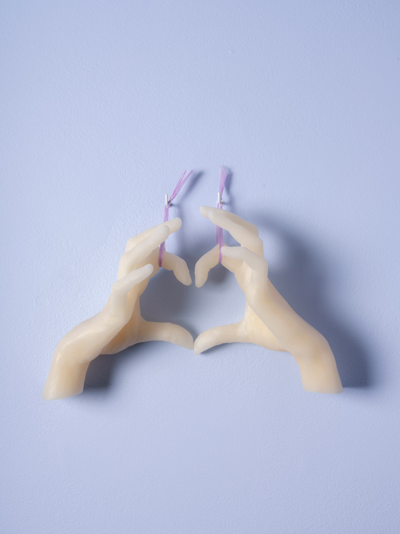Fragment de l'installation Spleen Spring. Moulage d'une paire de mains en cire suspendu par deux bolducs mauves, présenté sur fond bleu. Mains positionner de manière à former un cœur avec les doigts.