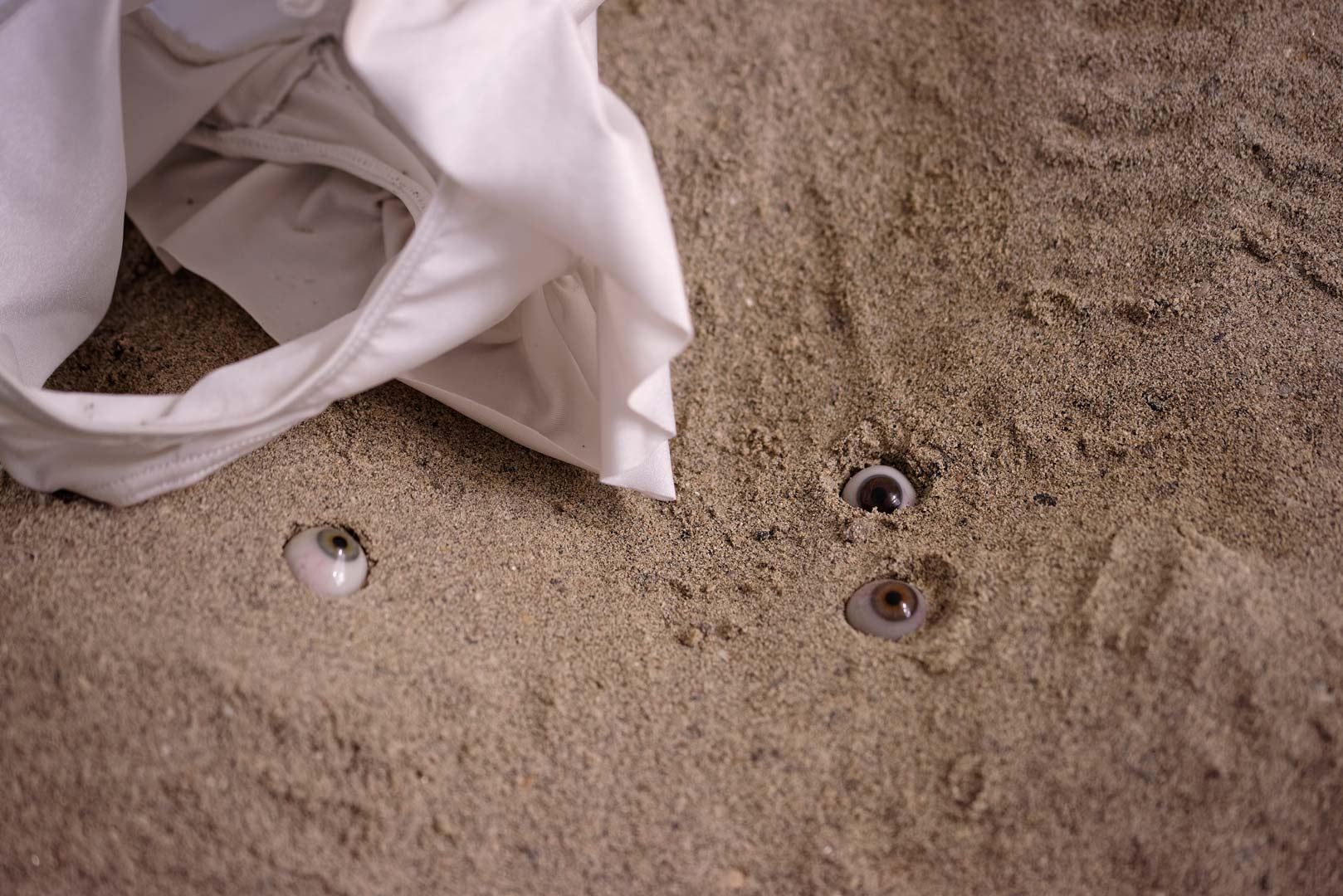 Vue de l'installation intitulée " Les tombeaux innocents". Focus resserré sur le sable. Des yeux de verre de différentes couleurs hésitent entre surgissement et enlisement. Ils semblent regarder dans différentes direction. A gauche, un morceau de maillot bain semble occupé mais pourtant vide.