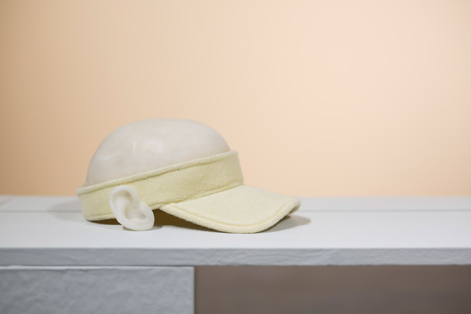 Fragment de l'installation Summer Seum. Focus sur un semblant de casquette en matière composites. Le chapeau est composé à partir d'une visière en éponge jaune pale auquel sont greffé une oreille de cire et une calotte en cire rappelant un crâne. La composition marque une hésitation entre casquette et crâne à nu.
