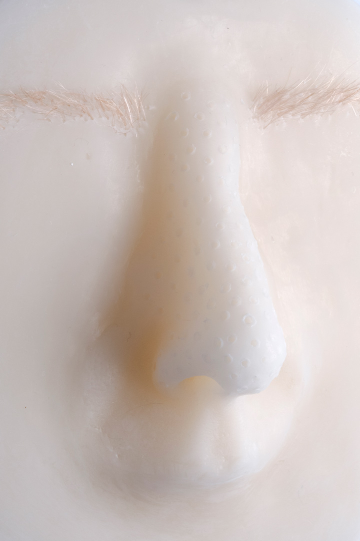 Zoom sur forme ovoïdale en cire avec nez et sourcils, permettant de découvrir des reliefs ressemblant à des pores dilatés.