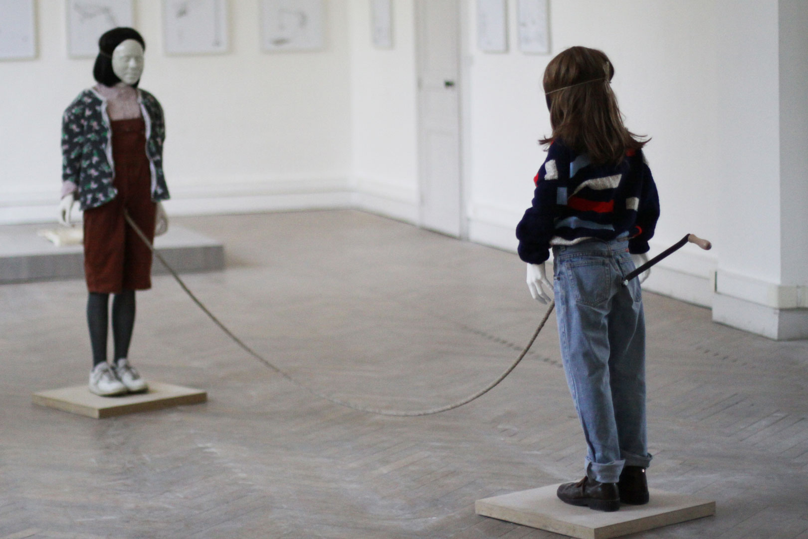 Vue d'ensemble de l'installation intitulée "Mouvement perpétuel". L'installation montre deux figures enfantines en face à face . Elles sont réunies par une corde sortant de leurs braguettes . La corde semble défier les lois de la gravité. Une manivelle sort de leur derrière.