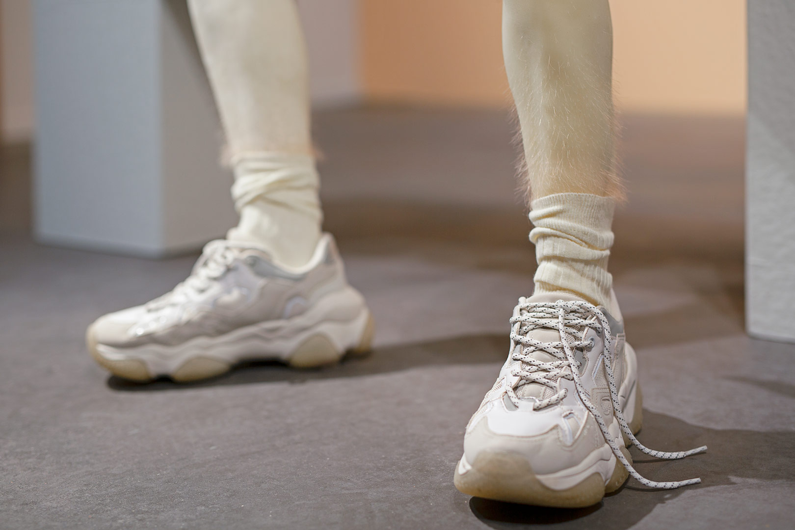 Fragment de l'installation Summer Seum. Focus resserré sur le bas d'une paire de jambes en cire portant des baskets et chaussettes beiges. Les mollets sont recouverts d'une fine pilosité rousses.