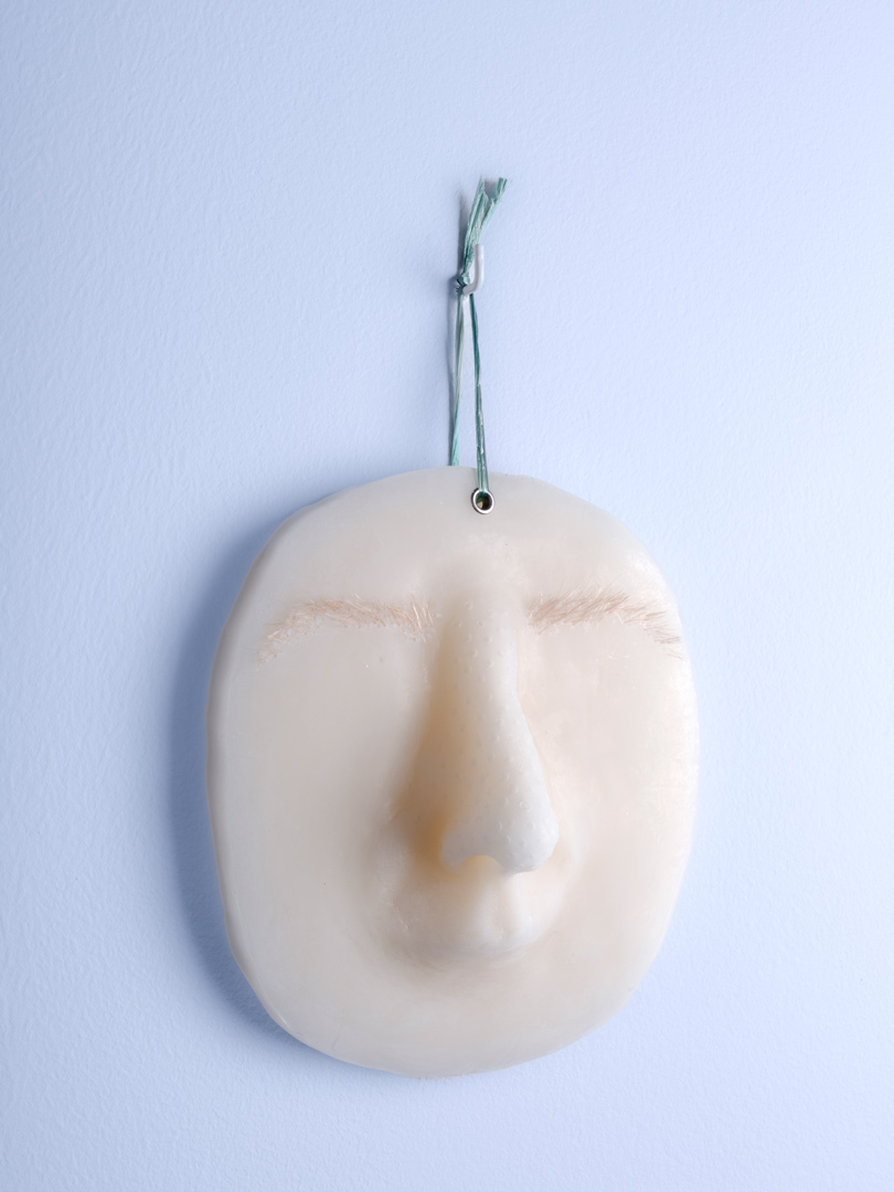 Fragment de l'installation Spleen Spring. Forme ovoïdale en cire avec nez et sourcils, suspendue par un bolduc vert. Fond bleu.