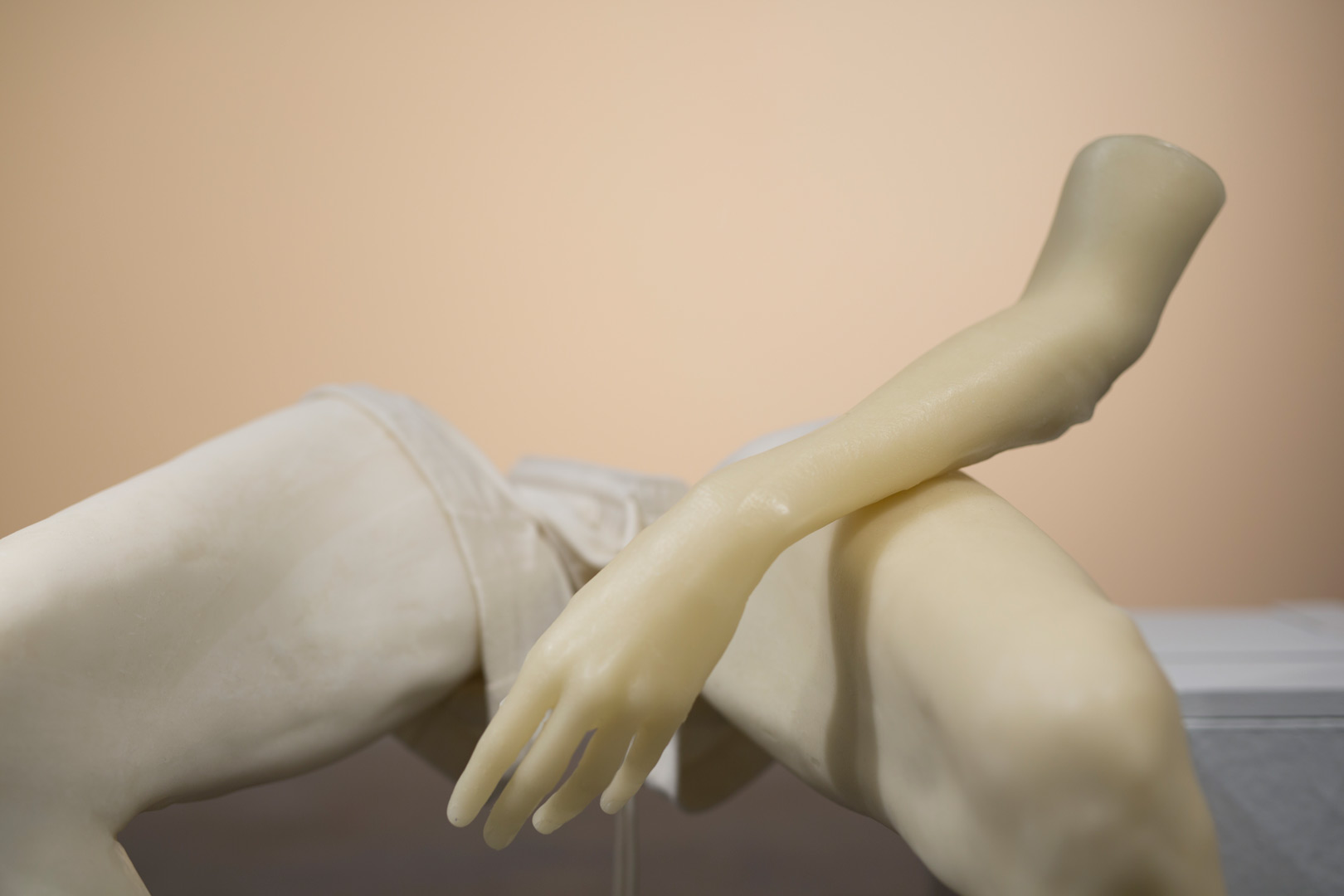 Fragment de l'installation Summer Seum, laissant apparaître un corps de cire morcelé figuré par deux jambes habillées d'un short en éponge beige à demi vide. Un avant bras est en appui sur la cuisse droite, il semble léviter.